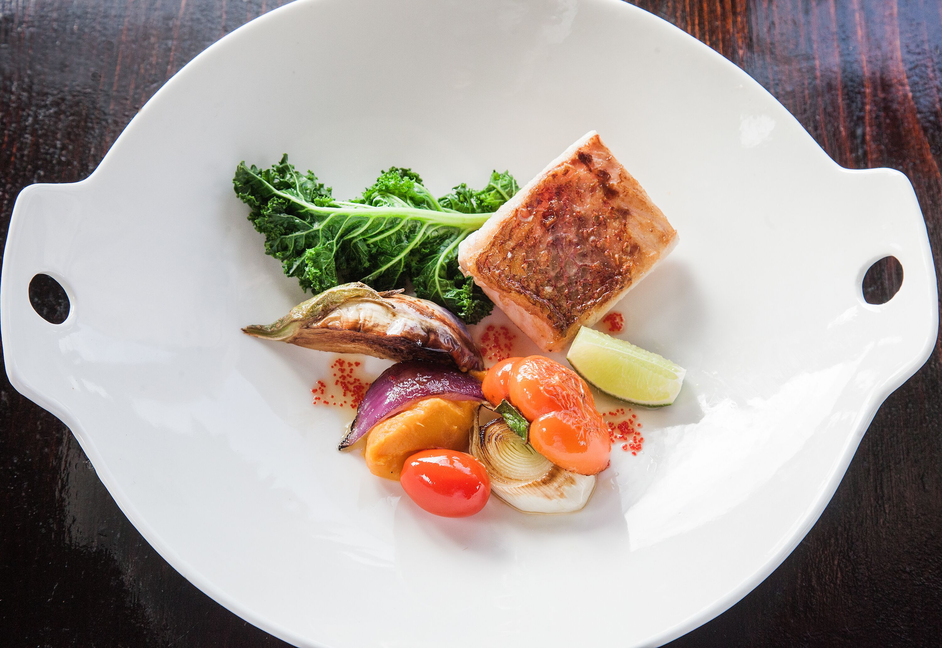 Une assiette blanche contenant divers aliments, notamment un morceau de viande grillée, des légumes et une tranche de citron vert.