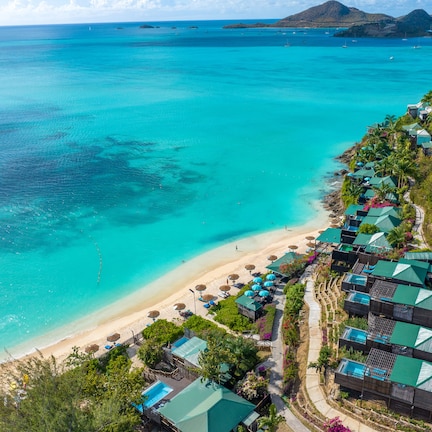 An aerial view of a coastal resort with a beach, clear turquoise waters, and a row of green-roofed villas.