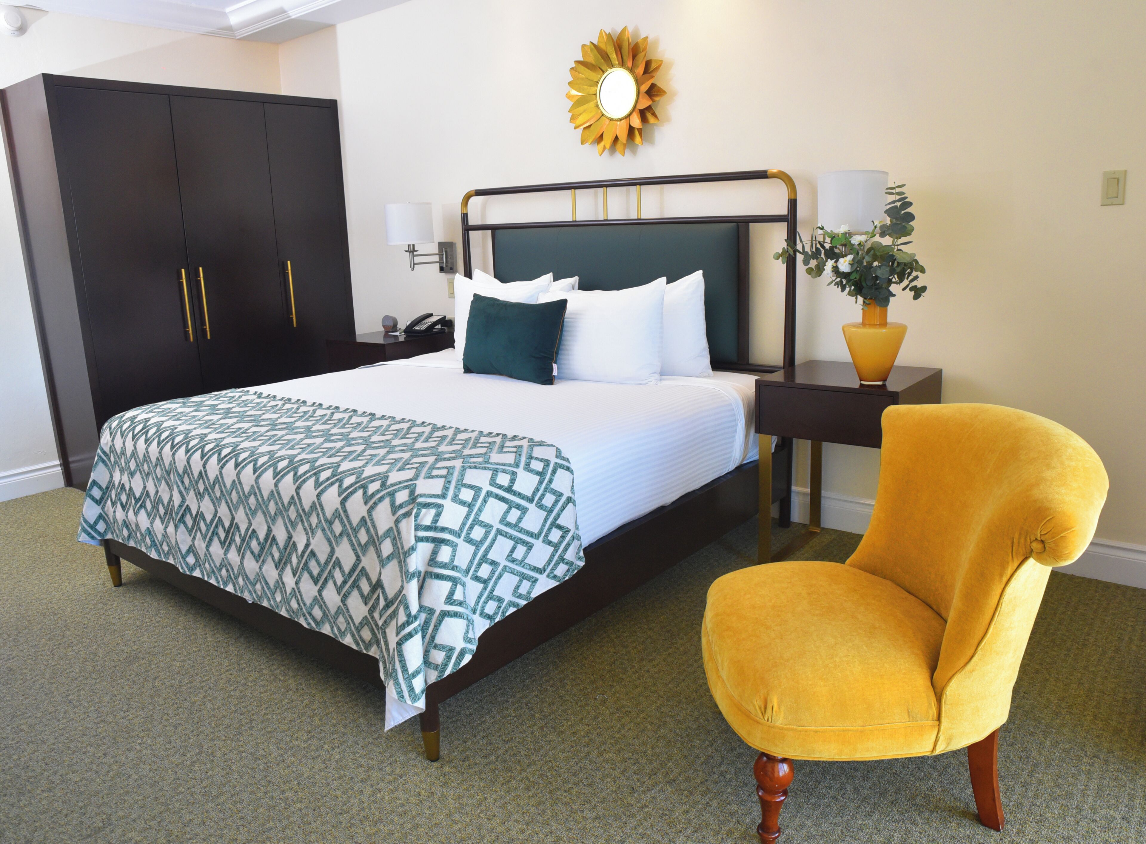 A hotel room with a bed, a nightstand, a chair, a wardrobe, and a sun-shaped wall decoration.