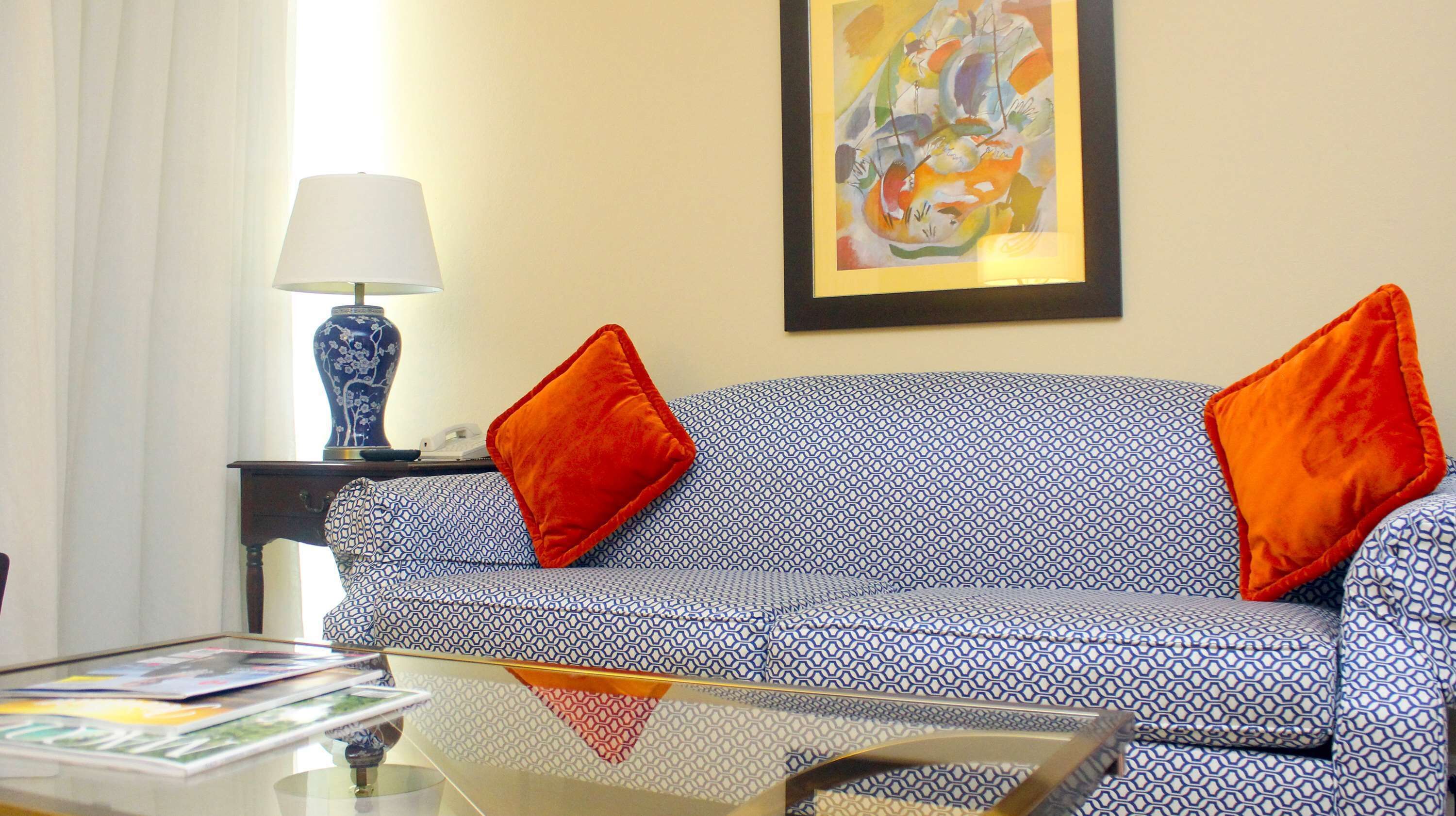 Un salon avec un canapé à motifs, deux coussins orange, un vase bleu et un tableau abstrait encadré accroché au mur.