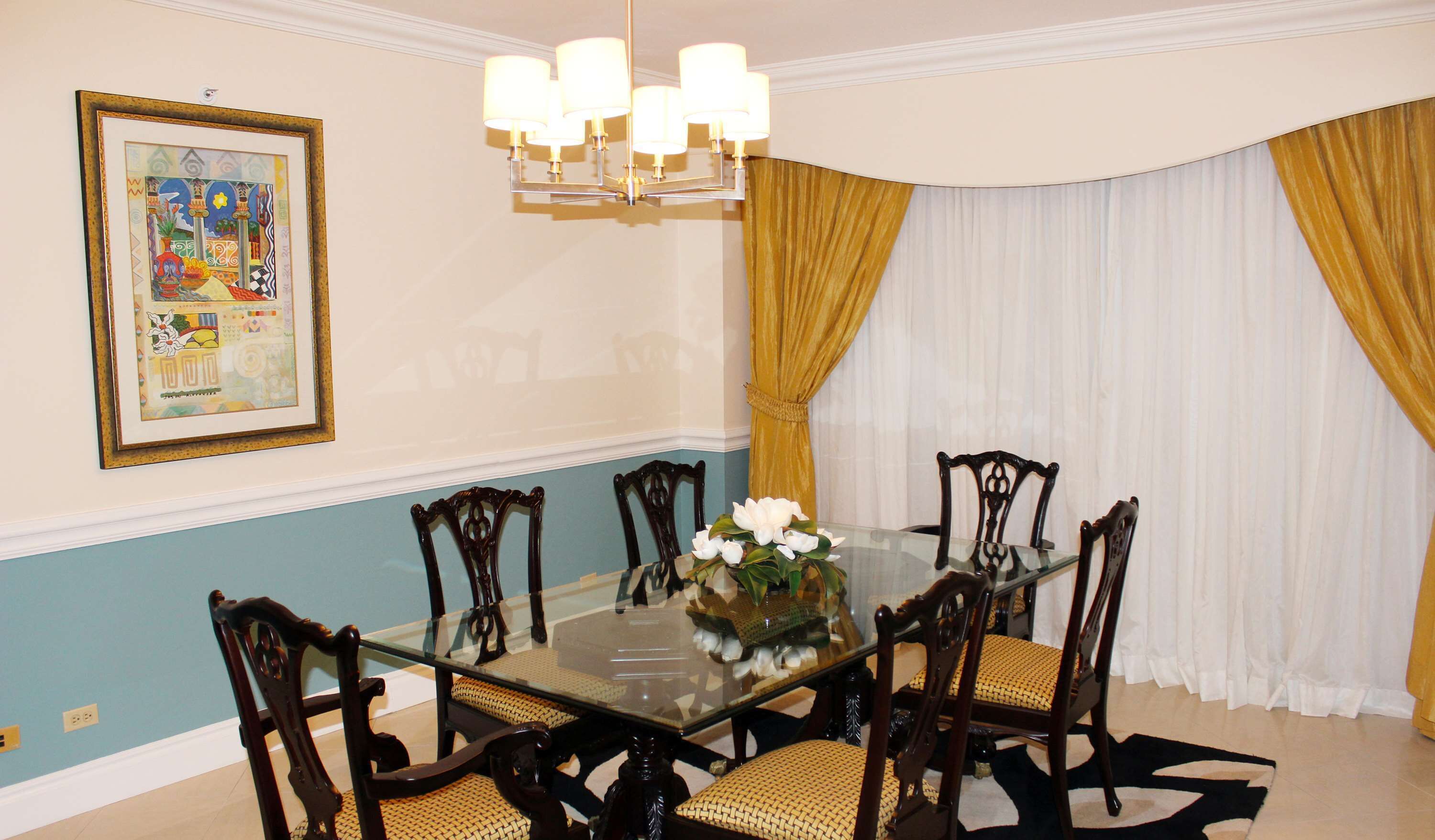 Een eetkamer met een glazen tafel, omringd door zes houten stoelen met geweven zittingen, een kroonluchter, een lijstwerk schilderij aan de muur, en een raam met gele gordijnen.