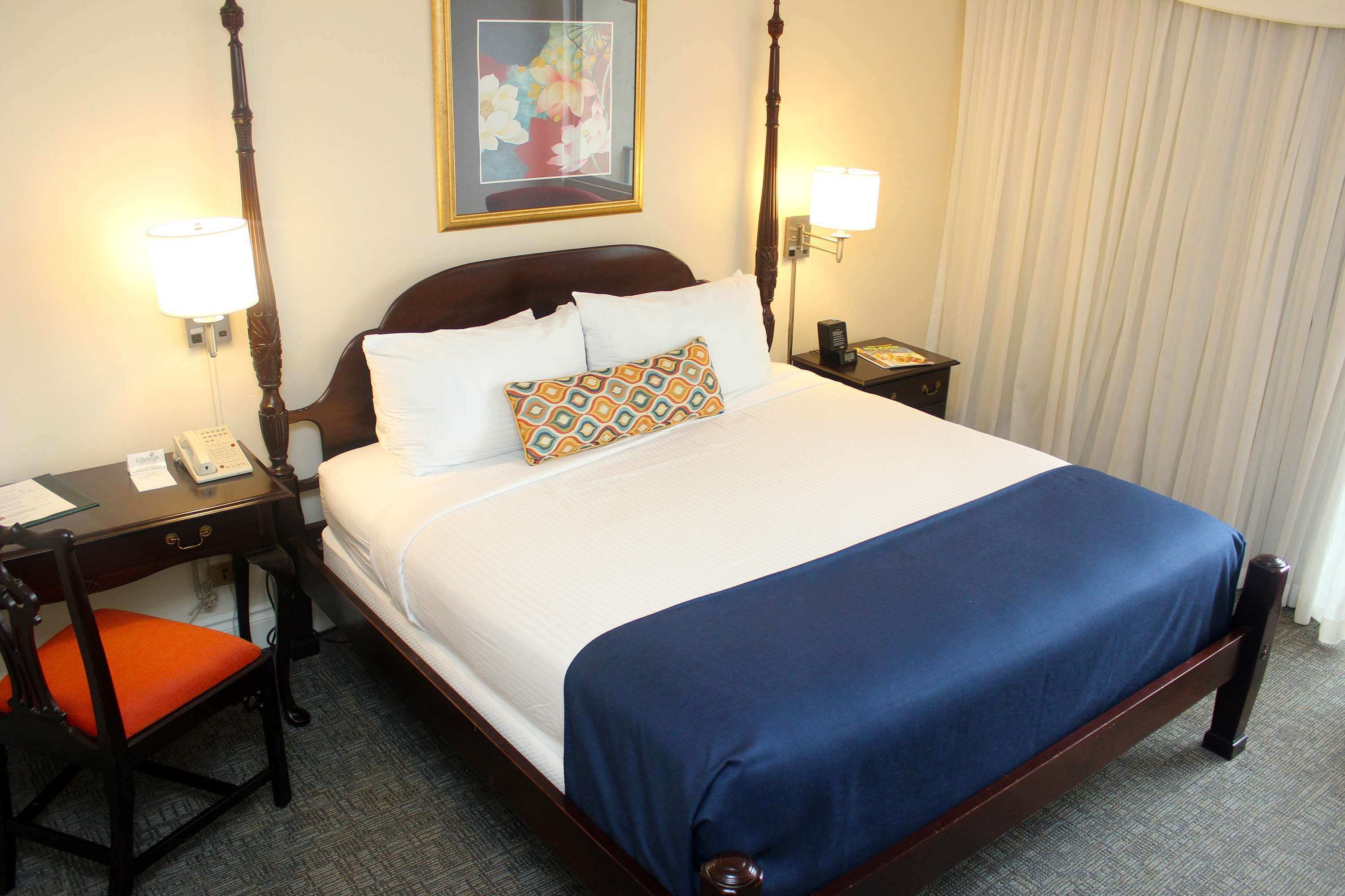 Quarto de hotel com uma cama grande, uma mesa de cabeceira de madeira com uma luminária, uma escrivaninha com um telefone e uma cadeira com almofada laranja.