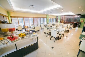 A spacious dining area with multiple tables and chairs, a buffet counter with various food items, and large windows with blinds. - The Courtleigh Hotel and Suites (Kingston)