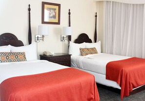 A hotel room with two beds, each with a white bedspread and red throw blankets. There are two wall-mounted lamps, a framed picture on the wall, and a window with white curtains. - The Courtleigh Hotel and Suites (Kingston)