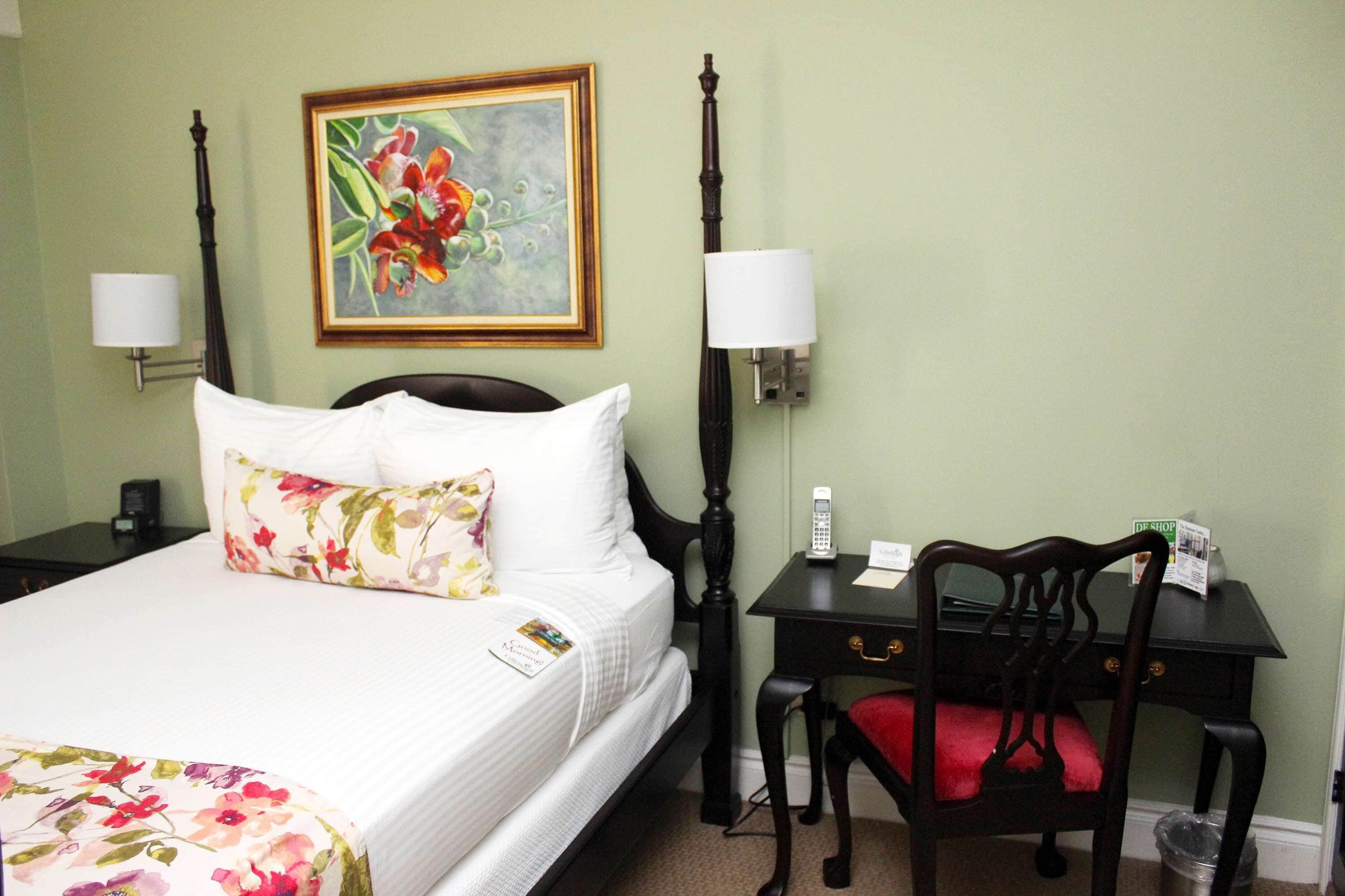 A hotel room with a bed, a desk, a chair, a lamp, a painting, and a trash can.
