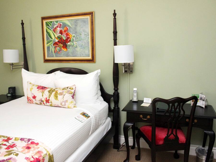 A hotel room with a bed, a desk, a chair, a lamp, a painting, and a trash can.
