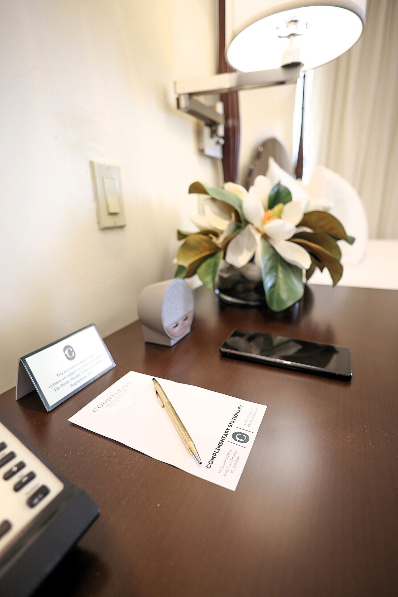 Escritorio de habitación de hotel con una tarjeta de presentación, un bolígrafo, un teléfono inteligente, una lámpara y un florero.