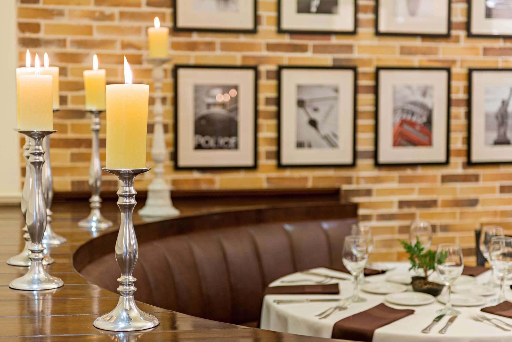 Un ambiente de restaurante con pared de ladrillo, cuadros enmarcados y velas sobre pedestales de plata.