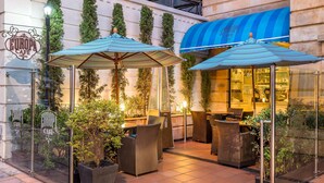 Un área de café al aire libre con asientos y sombrillas, rodeada de follaje y una fachada de edificio con toldo azul.