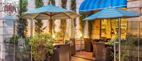 An outdoor cafe area with seating and umbrellas, surrounded by greenery and a building facade with a blue awning.