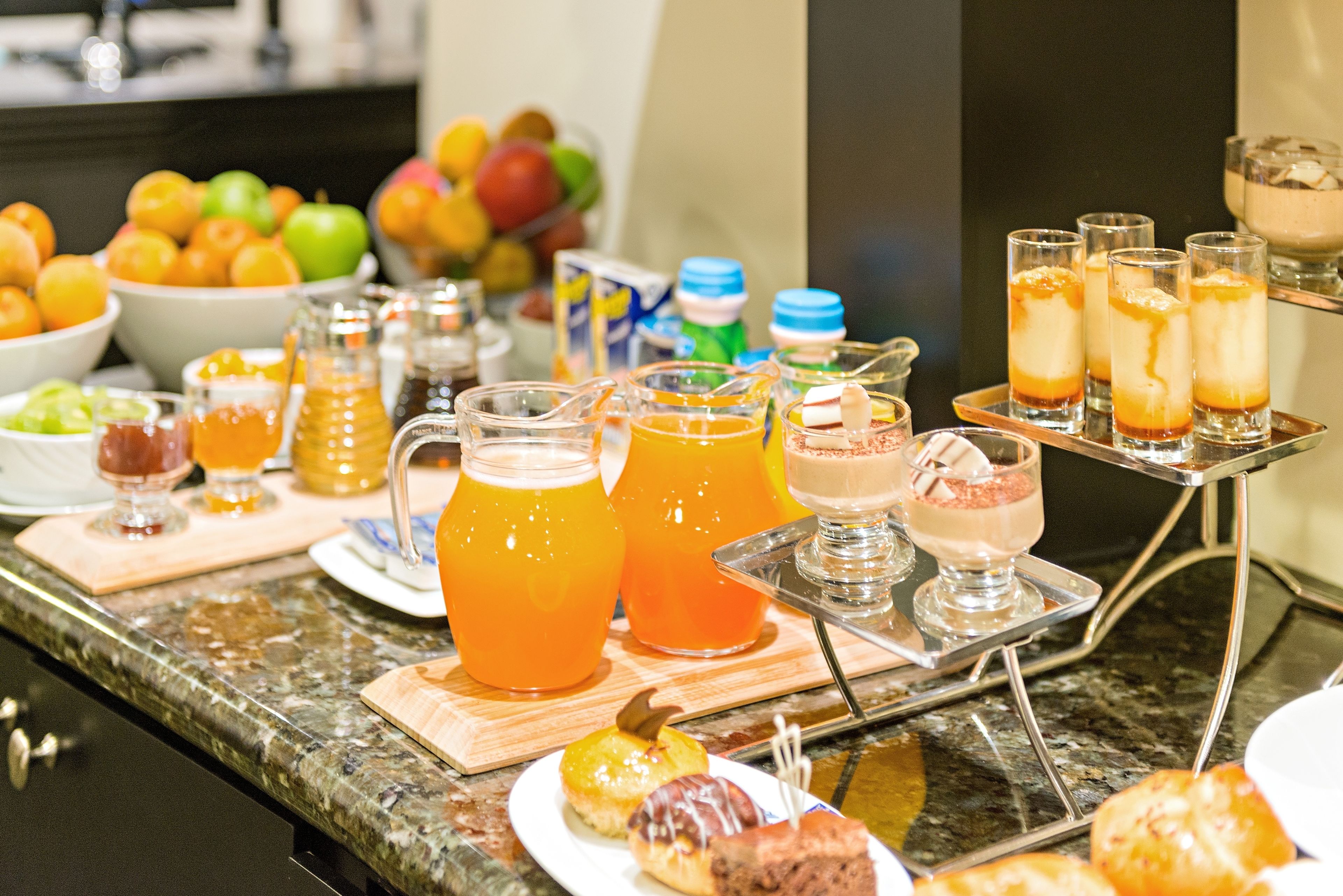 Un bufé con diversos alimentos y bebidas, que incluye un bol de fruta, una jarra de jugo de naranja y varias opciones de postres.