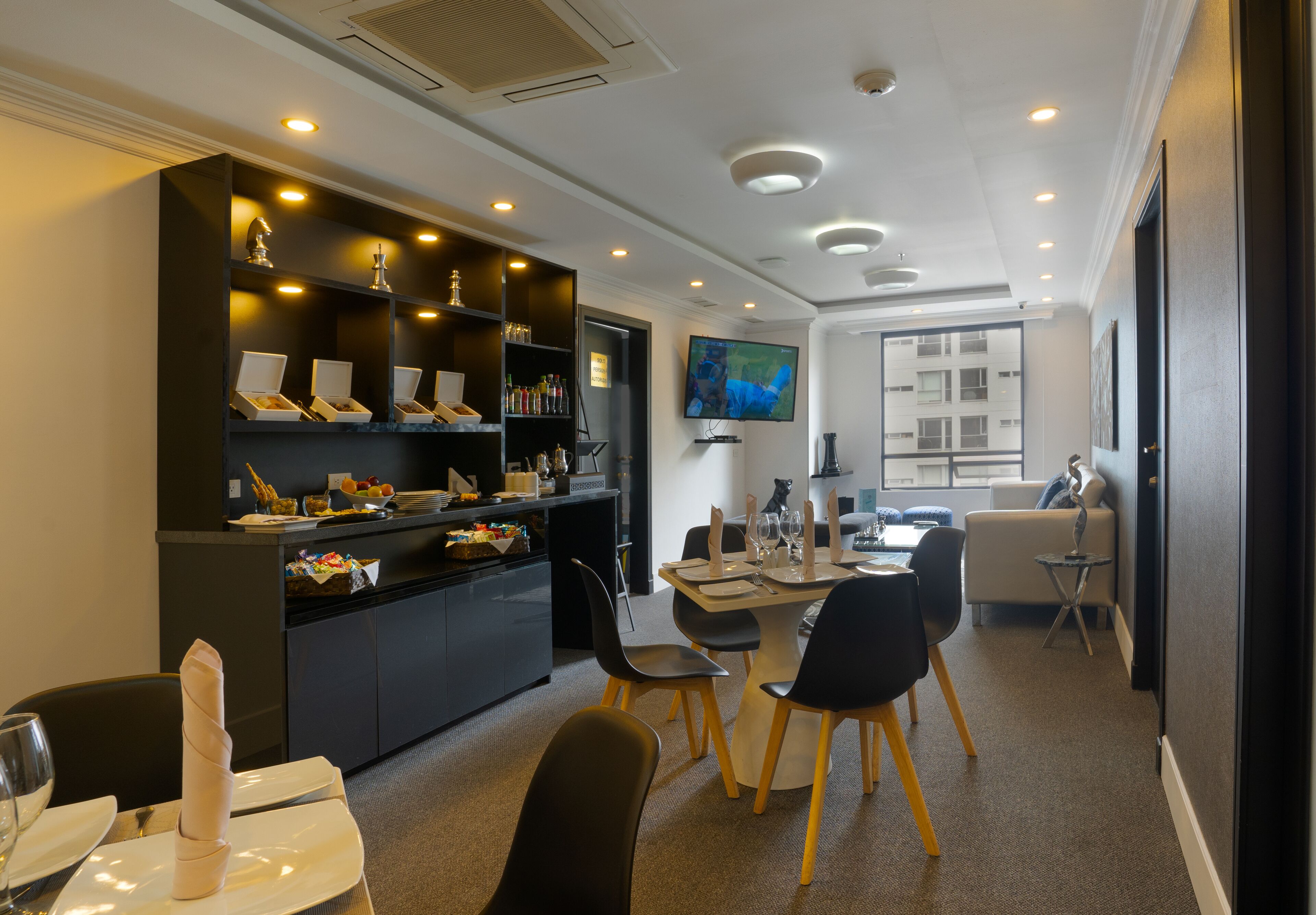A modern dining area with a central table set for four, surrounded by black chairs with wooden legs. A black cabinet with shelves on the left holds various items, and a flat-screen TV is mounted on the wall above the table.