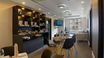 A modern dining area with a central table set for four, surrounded by black chairs with wooden legs. A black cabinet with shelves on the left holds various items, and a flat-screen TV is mounted on the wall above the table.