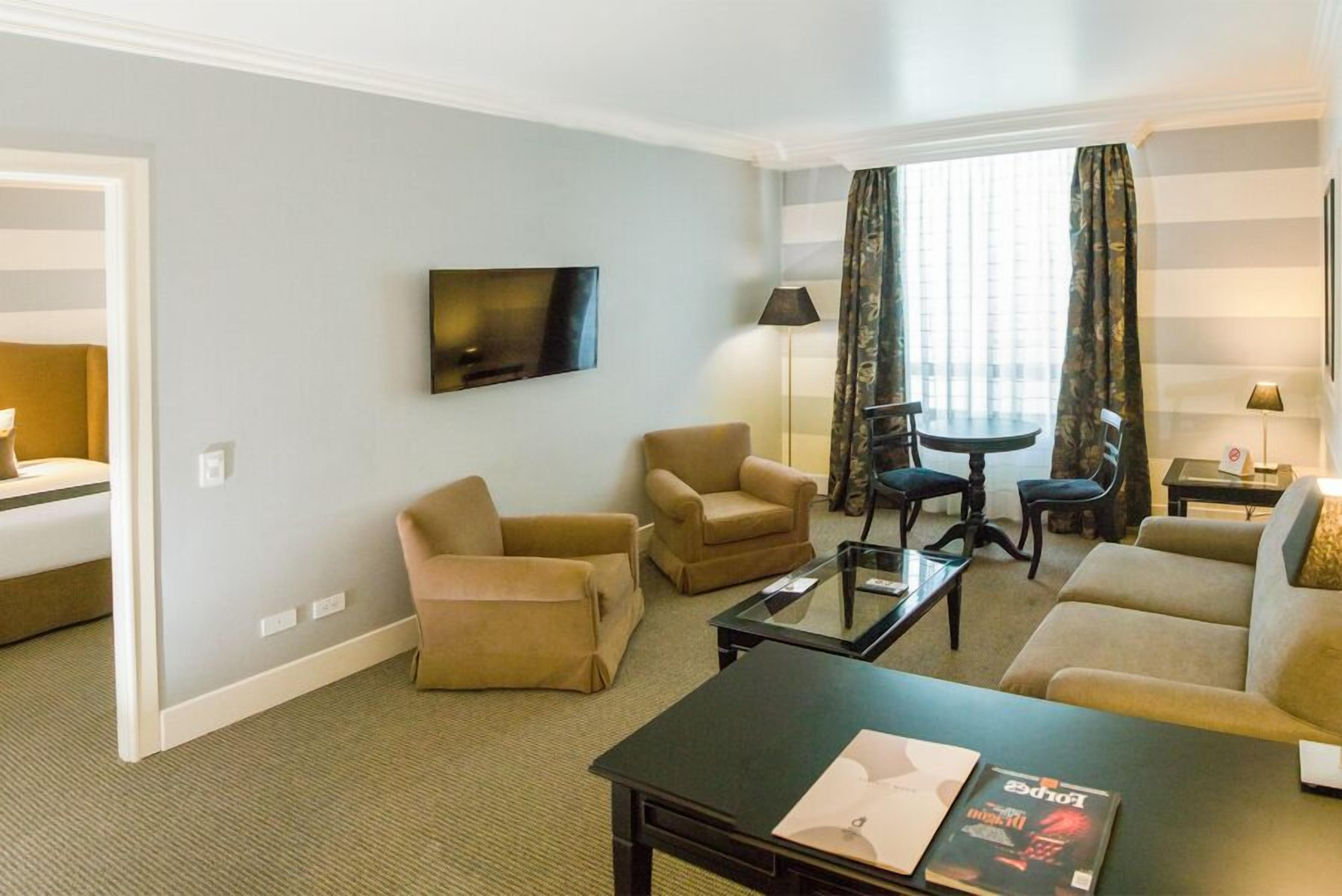 Une chambre d’hôtel avec un canapé, deux fauteuils, une table basse, un téléviseur, une lampe et une fenêtre avec des rideaux.
