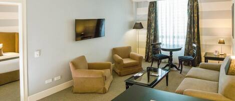 Habitación de hotel con sofá, dos sillones, una mesa de centro, televisor, lámpara y ventana con cortinas.