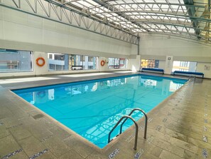 Una piscina interior con agua azul cristalina, suelo de baldosas y estructura de techo metálica.