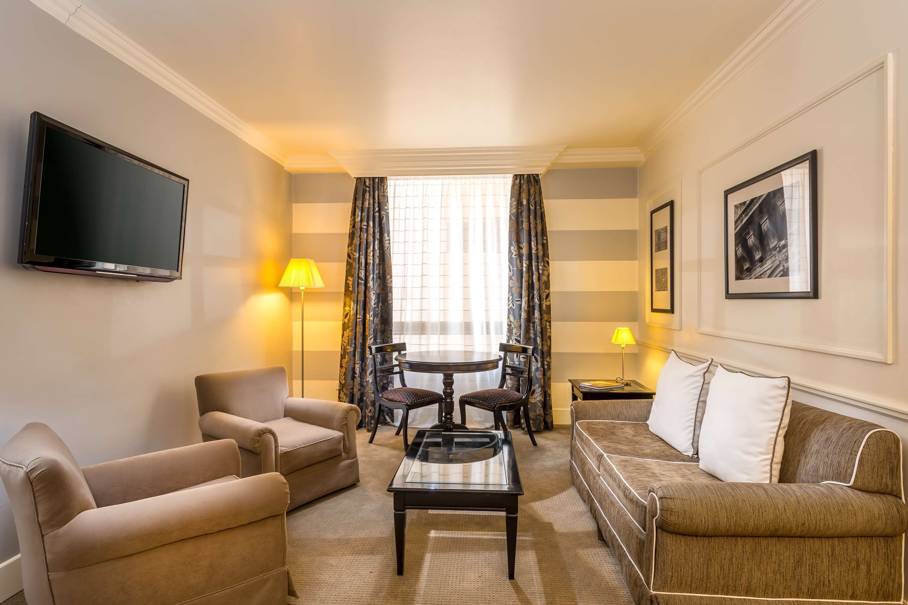 A hotel room with a flat-screen TV, two sofas, a coffee table, a dining table with chairs, and a window with curtains.