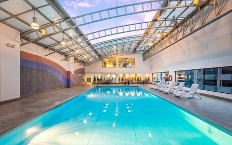 An indoor swimming pool with a glass ceiling, lounge chairs, and a view of the cityscape.