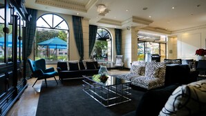 Un hall amplio con un sofá moderno, un sillón azul y una mesa de centro. Ventanas grandes con cortinas ofrecen luz natural y vistas al exterior.