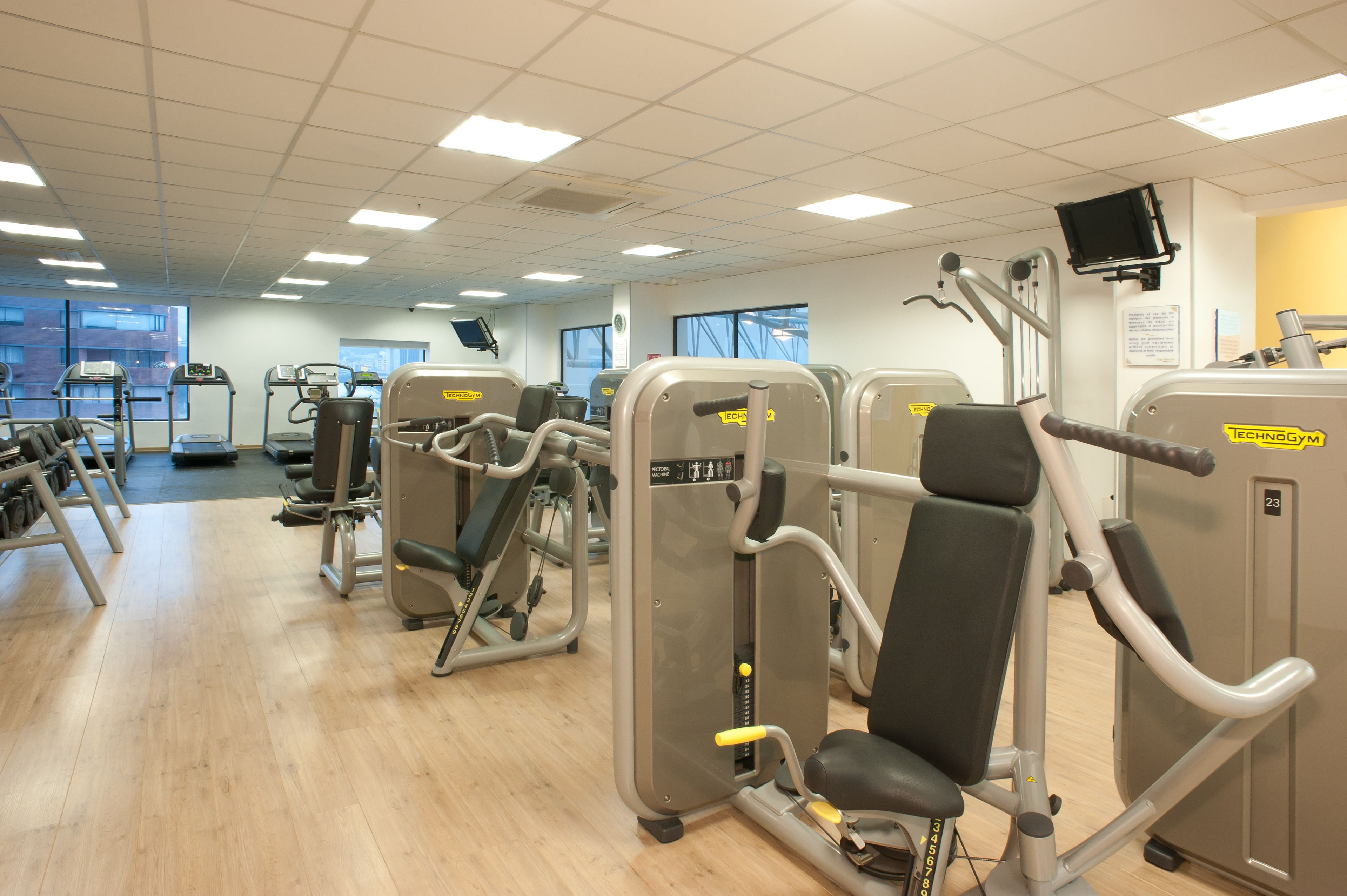 A gym with various exercise machines, large windows, and a clean, organized layout.