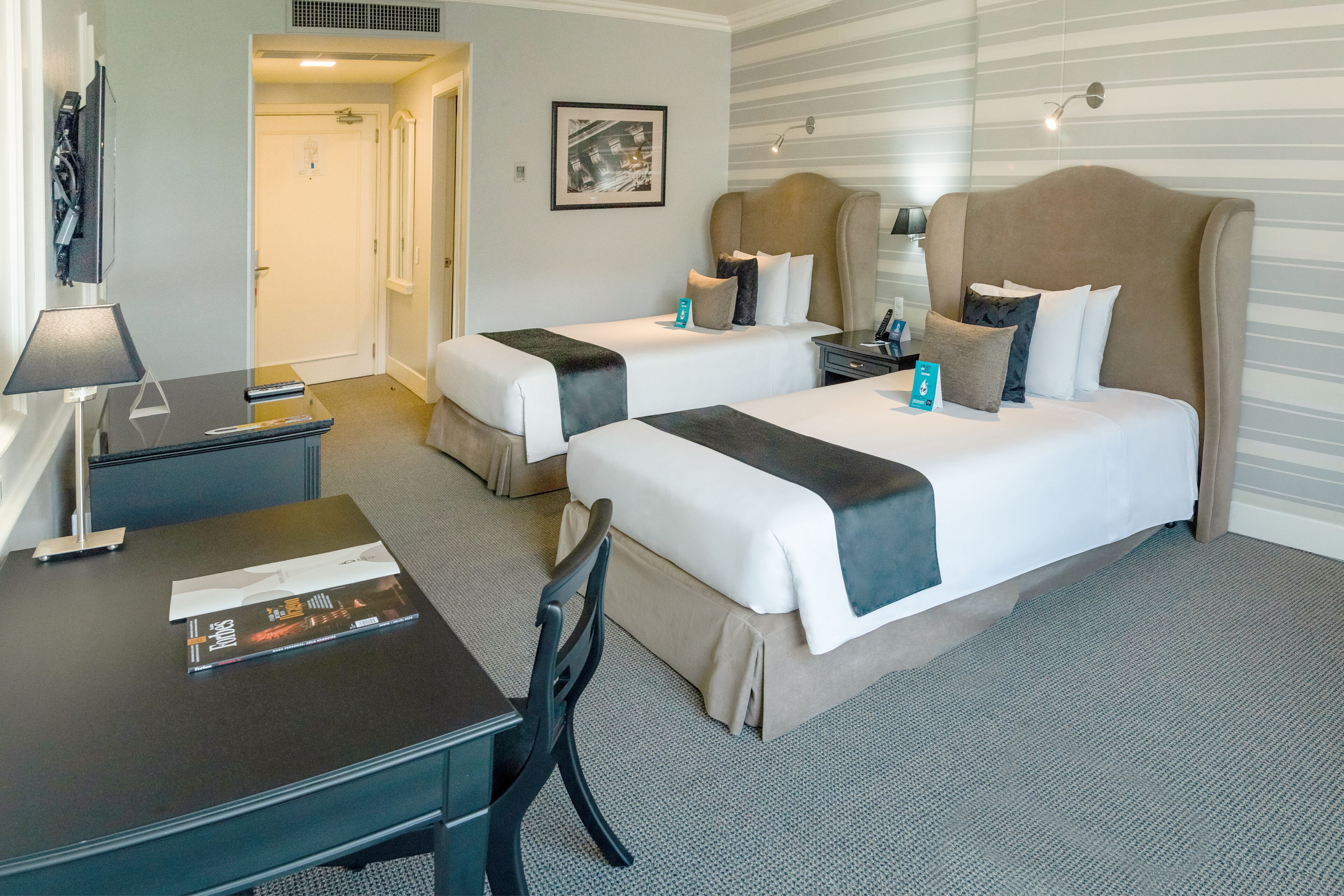 A hotel room with two beds, a desk, a chair, a TV, and a framed picture on the wall.