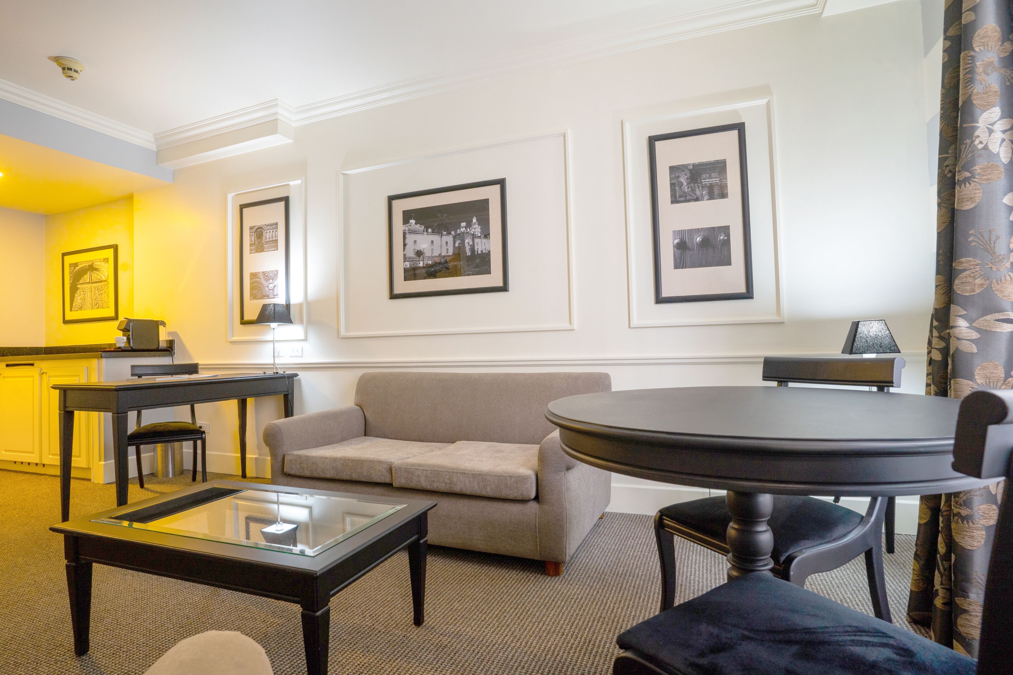 A hotel room with a sofa, a glass-top coffee table, a round dining table, and framed pictures on the wall.