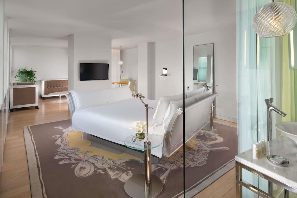Une chambre d’hôtel moderne dotée d’un grand lit, d’un coin salon avec un canapé et une petite table, d’un téléviseur à écran plat et d’une salle de bain fermée par des parois de verre.