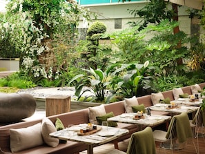 A restaurant with outdoor seating, surrounded by lush greenery and a variety of plants. Tables are set with plates, glasses, and cutlery.