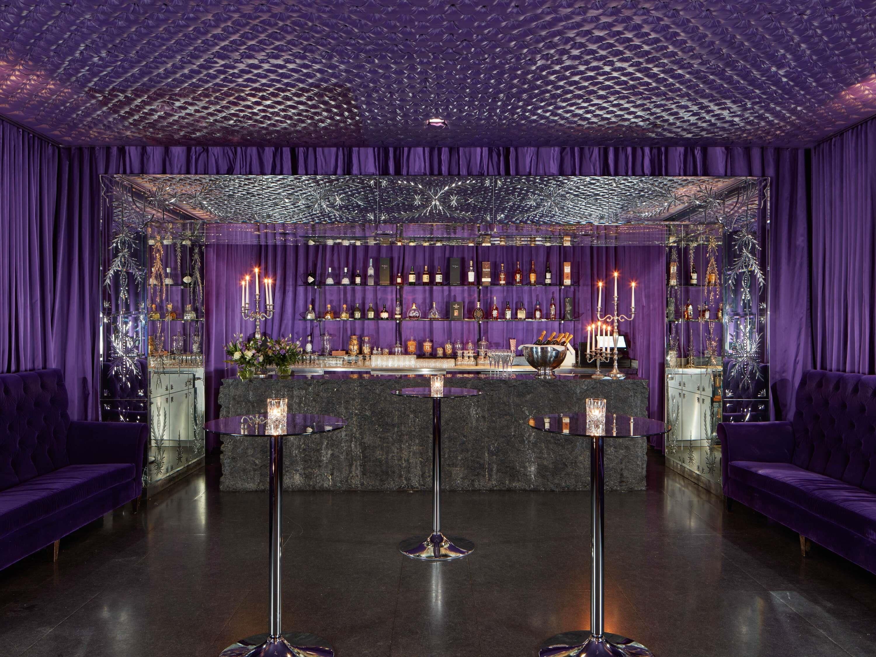 Elegante zona de bar con cortinas moradas, una barra central y dos taburetes.