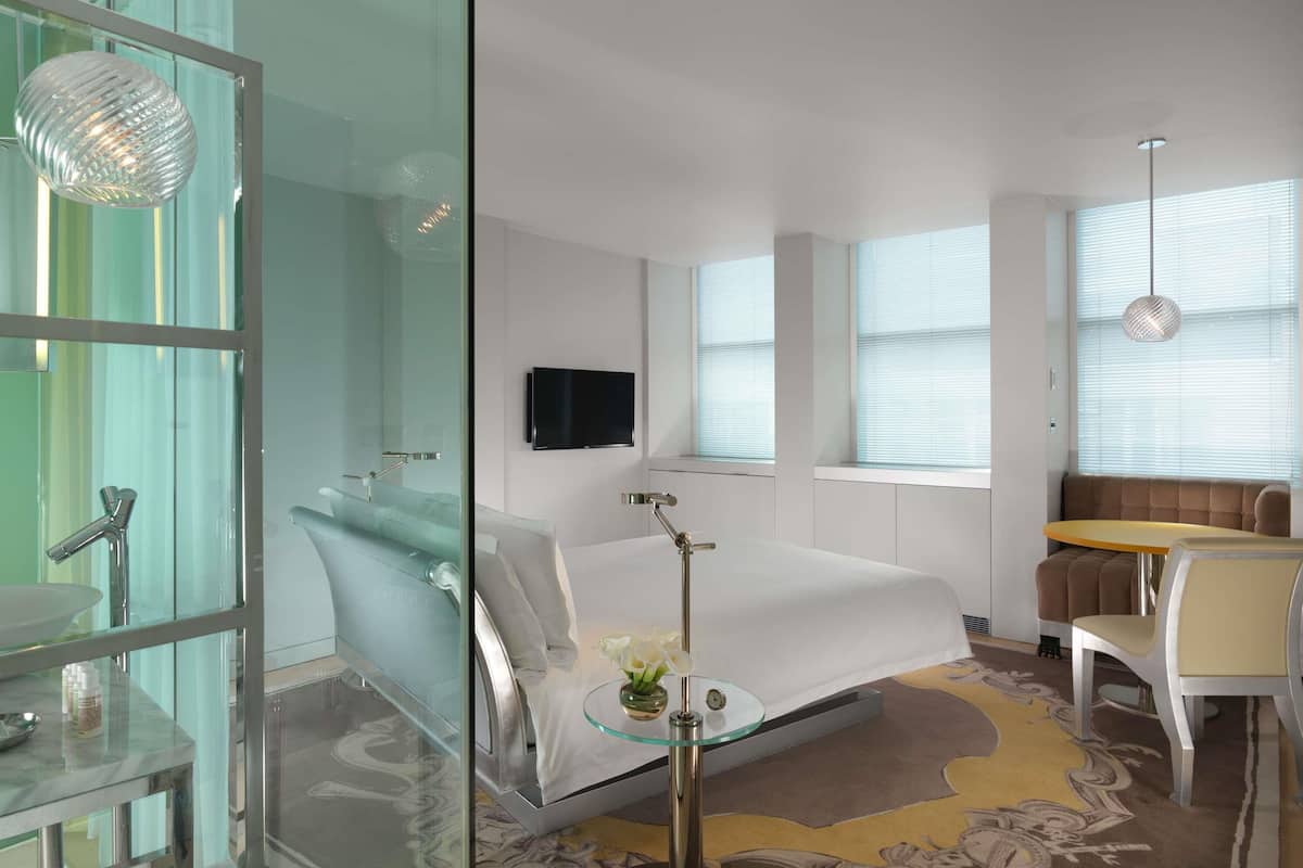 Une chambre d’hôtel moderne dotée d’une salle de bain entièrement vitrée, d’un lit avec une table de chevet en verre, d’une télévision et d’un coin salon avec une table et des chaises.