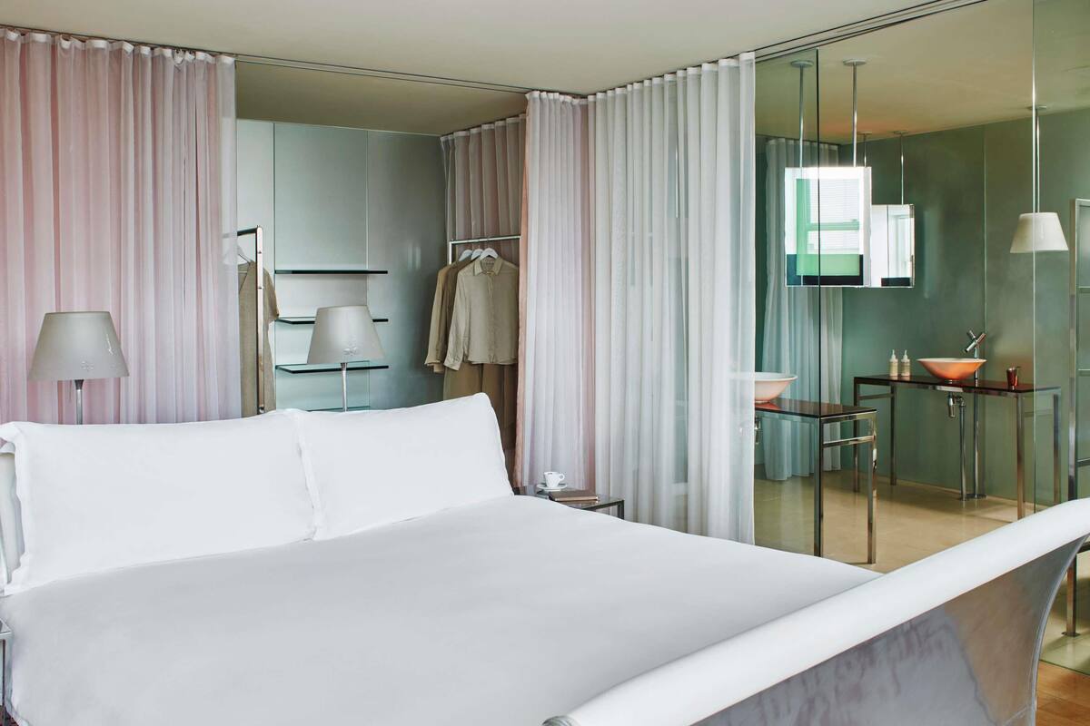 Une chambre d’hôtel moderne dotée d’un grand lit, d’une salle de bain entièrement vitrée et d’un coin salon avec une table et des chaises.