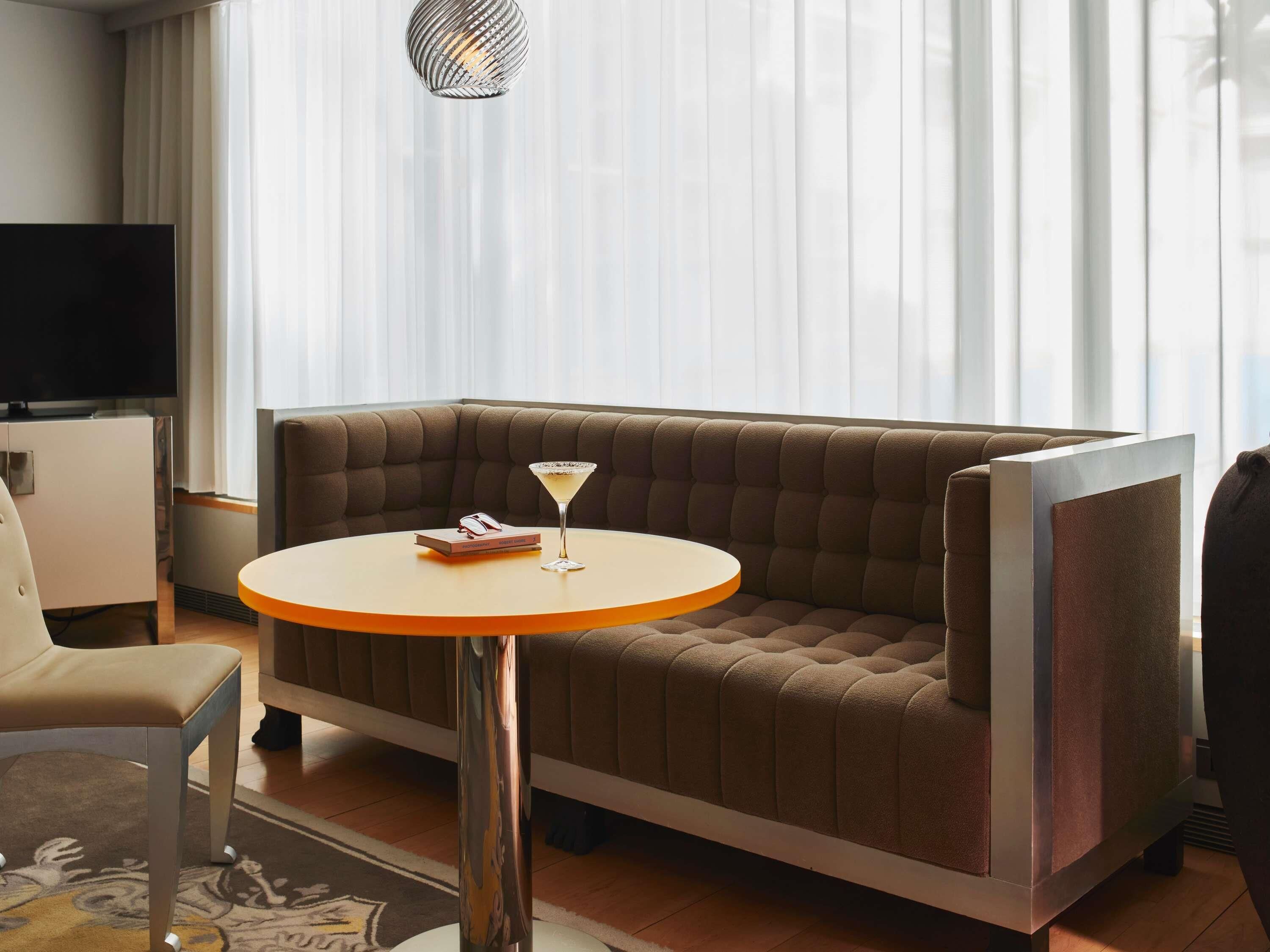 A modern hotel room with a brown upholstered sofa, a round table with a martini glass, and a television on a cabinet.
