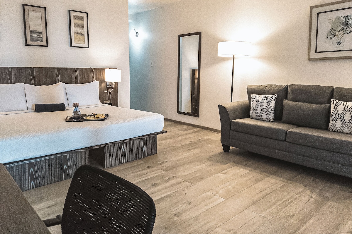 Une chambre d’hôtel avec un lit, un canapé, une chaise et une table à manger. Des tableaux encadrés ornent le mur et une lampe est posée sur le canapé.