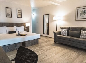 Habitación de hotel con cama, sofá, sillón y una mesa de comedor. En la pared hay cuadros enmarcados y una lámpara sobre el sofá.