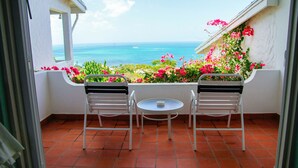 Un balcon avec deux chaises et une petite table, donnant sur un jardin où fleurissent des fleurs roses et offrant une vue sur l’océan.