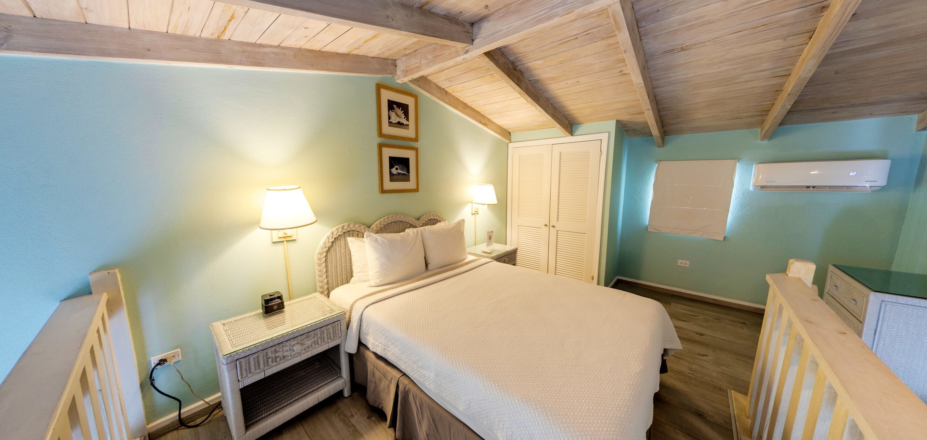 A bedroom with a bed, bedside lamps, a nightstand, a wall-mounted air conditioner, and a door with louvered shutters.
