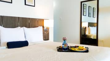 Habitación de hotel con cama, una bandeja de frutas y un jarrón de flores, y un espejo que refleja la habitación.