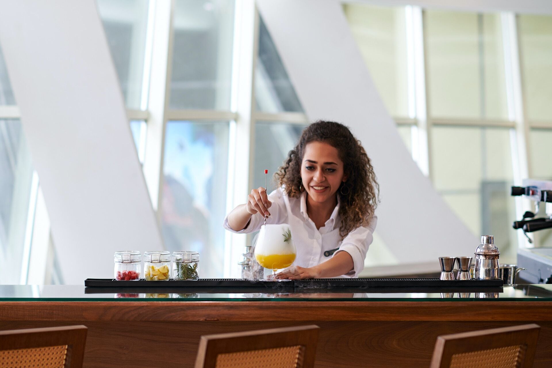 A person preparing a drink at a bar with a modern interior, including a glass counter and a large window.