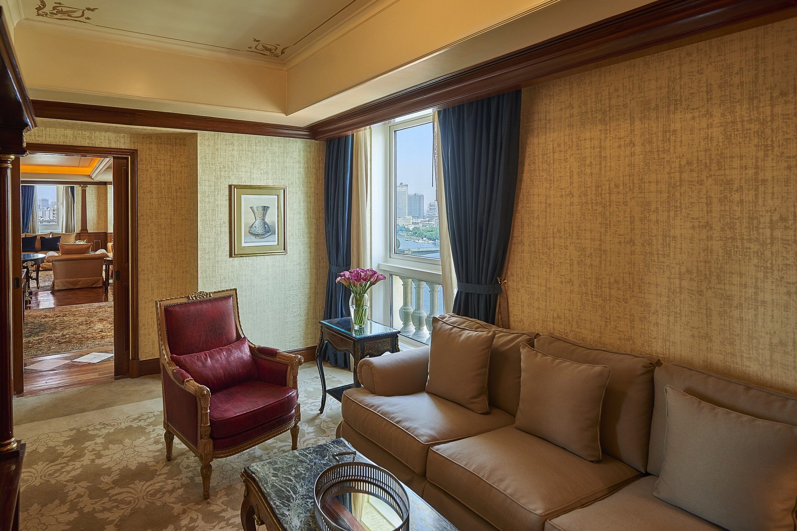 A hotel room with a red armchair, beige sofa, and a view of the city through large windows.