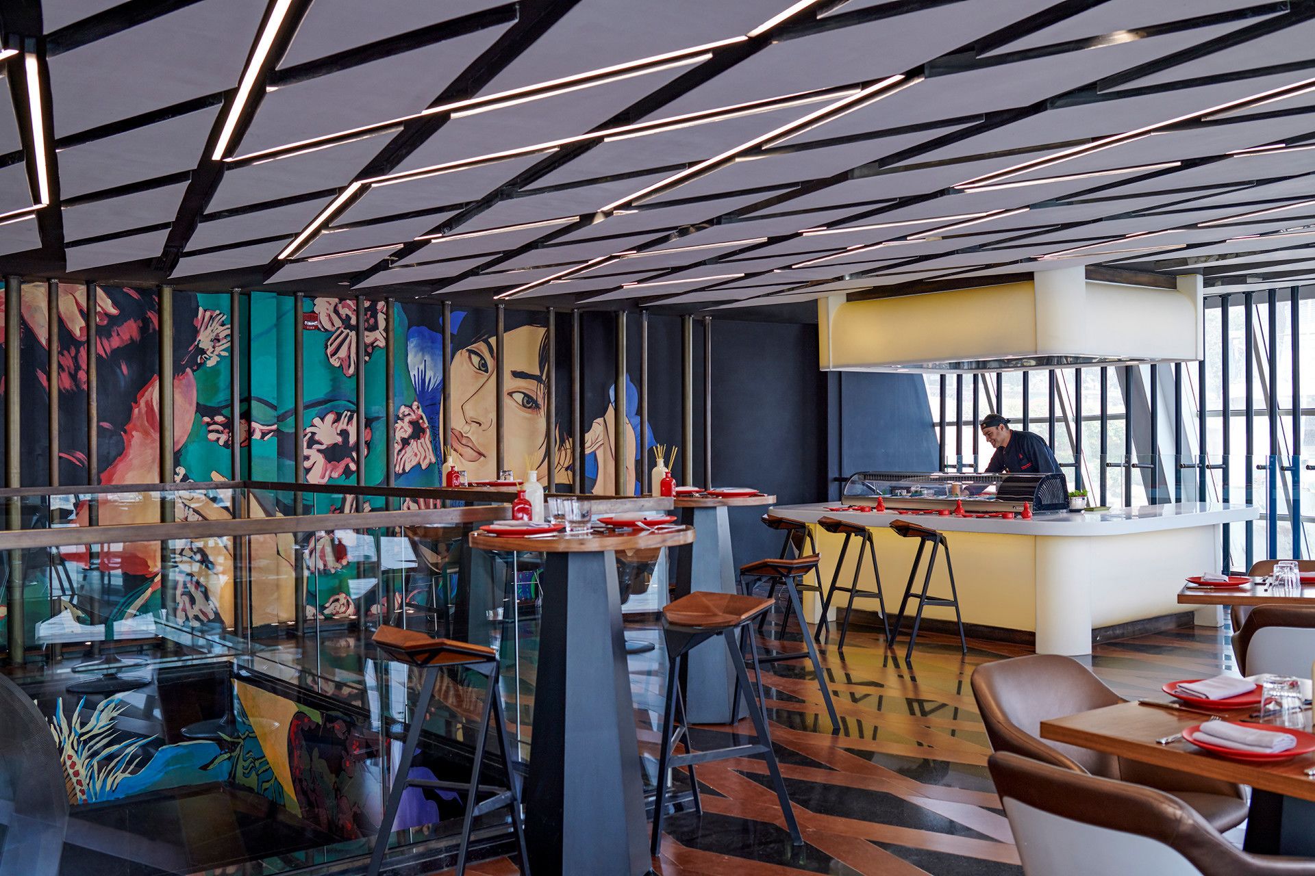 A modern restaurant with high ceilings, geometric ceiling design, and vibrant artwork on the walls.