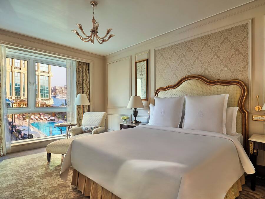 A spacious hotel room with a large bed, a sitting area with a view of a pool, and a chandelier.
