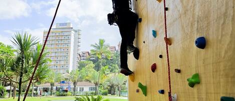 A person climbing a wooden rock wall with colorful holds, surrounded by a grassy area with palm trees and a building in the background.