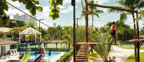 A rope bridge over a pool with slides, palm trees, and a gazebo.