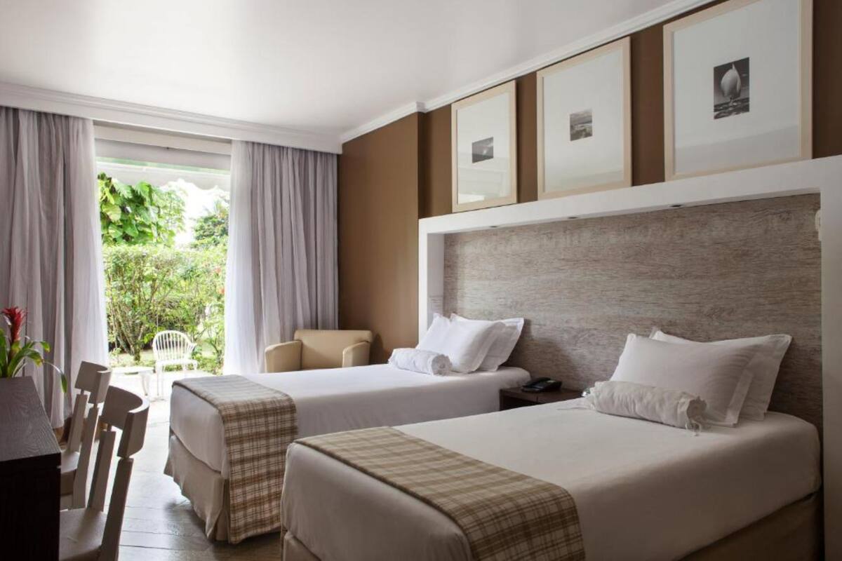 Habitación de hotel con dos camas, un escritorio, una silla y vistas al verde exterior.
