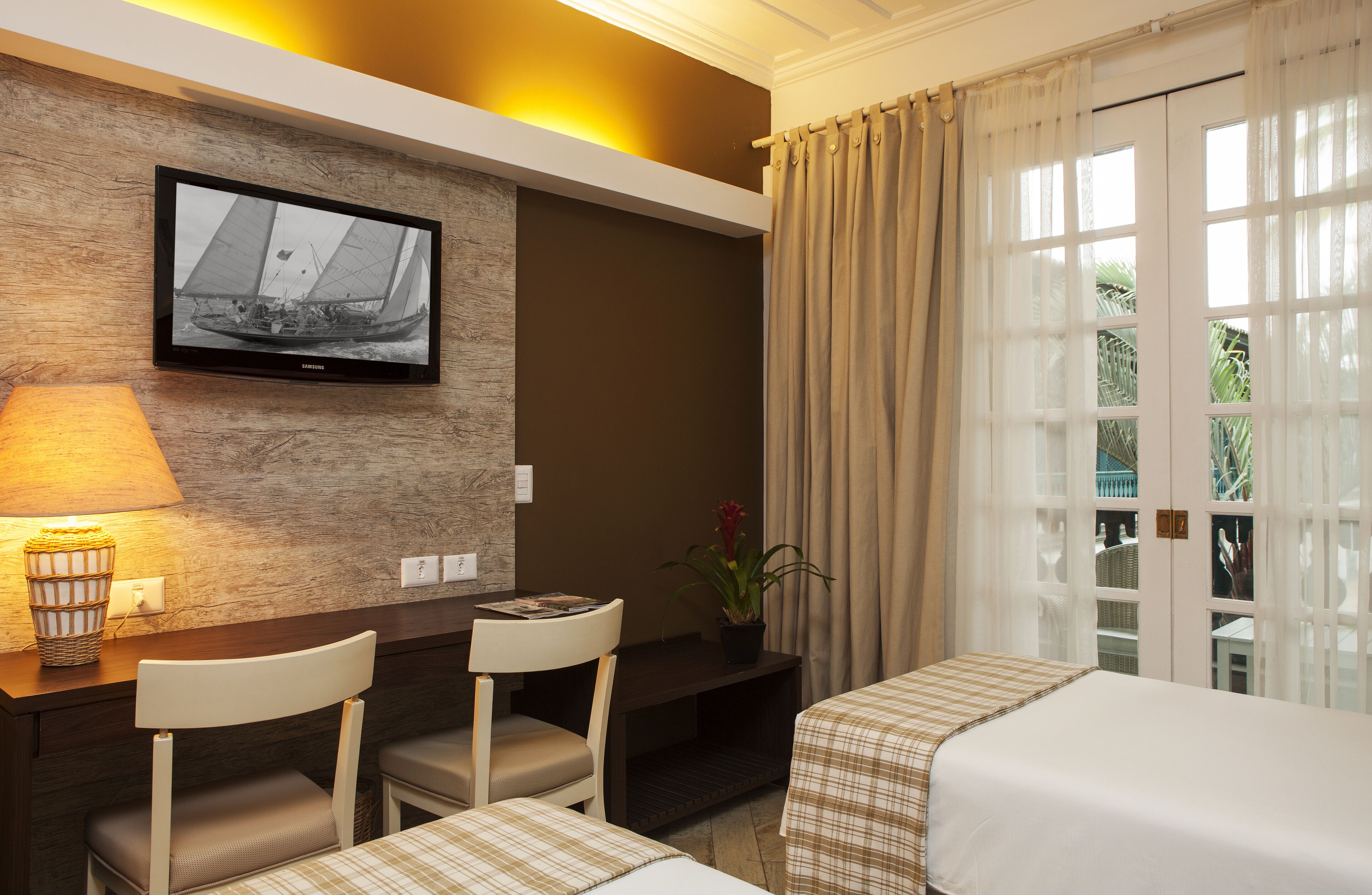 Habitación de hotel con televisión montada en la pared, escritorio con sillas, cama con cubrecama a cuadros y ventana con cortinas.