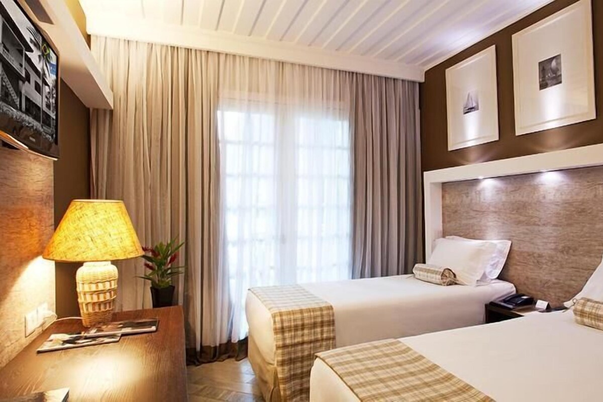 A hotel room with two beds, a nightstand, a lamp, a potted plant, and a window with curtains.