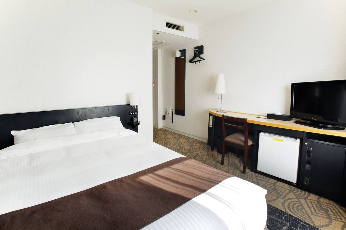 A hotel room with a bed, a desk with a computer, a TV, and a small fridge.
