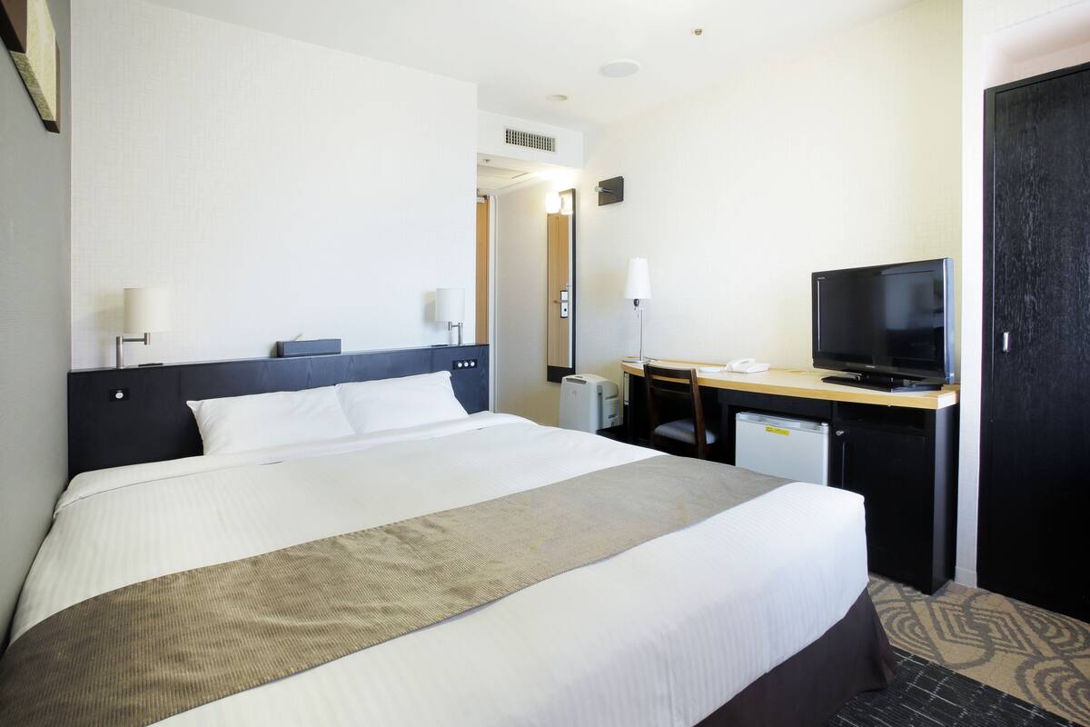 A hotel room with a large bed, a desk with a television, and a small kitchenette.