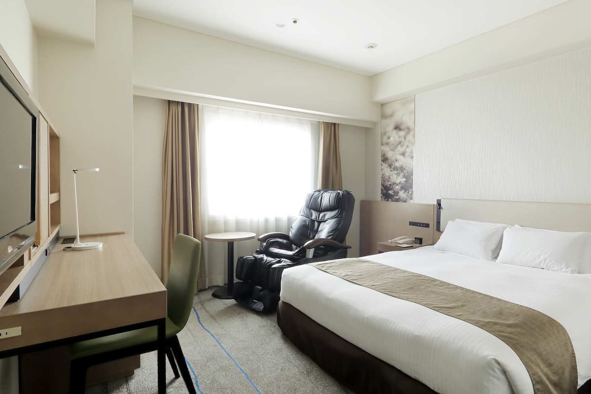 Une chambre d’hôtel équipée d’un lit, d’un bureau, d’une chaise, d’un fauteuil massant, d’une télévision et d’une fenêtre avec des rideaux.