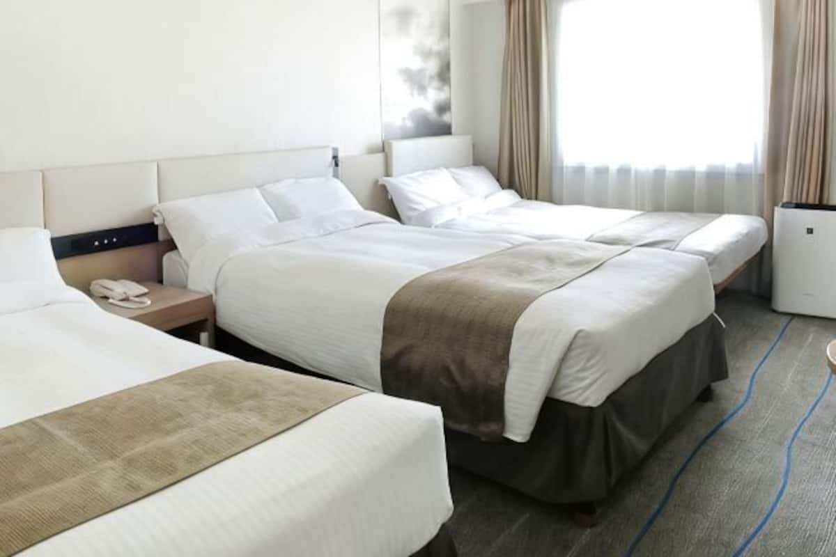 A hotel room with three beds, a bedside table with a phone, and a window with curtains.
